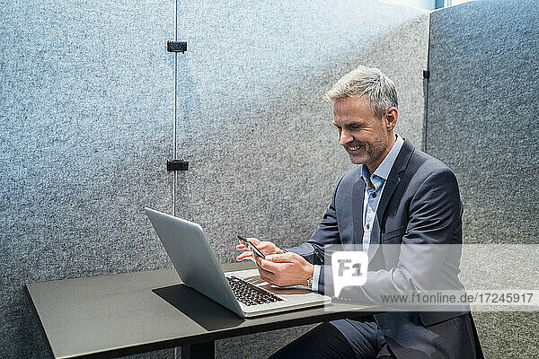 Smiling male entrepreneur with laptop using mobile phone in office