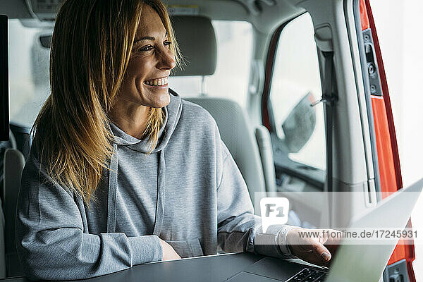 Fröhliche Unternehmerin mit Laptop  die wegschaut  während sie in einem Lieferwagen sitzt