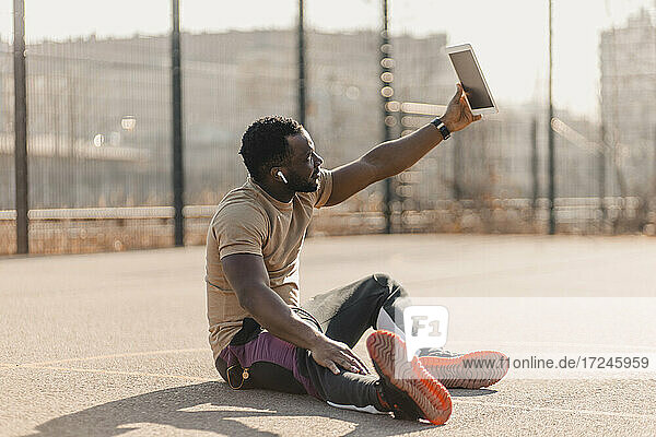 Junger Mann nimmt Selfie durch digitale Tablette beim Sitzen auf Sportplatz während sonnigen Tag