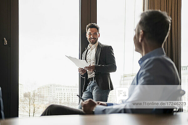 Lächelnder Geschäftsmann mit Dokument im Gespräch mit einem männlichen Kollegen am Arbeitsplatz