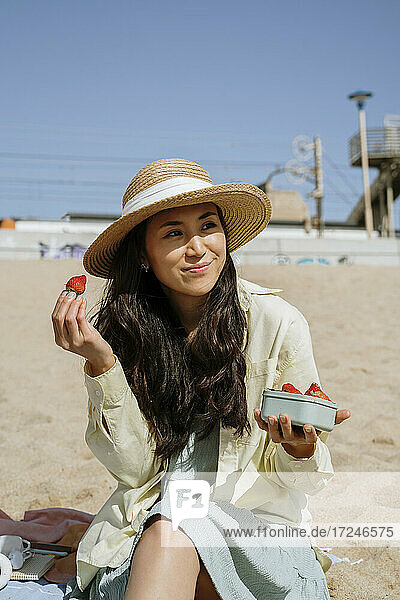 Frau hält Erdbeeren  während sie an einem sonnigen Tag am Strand sitzt