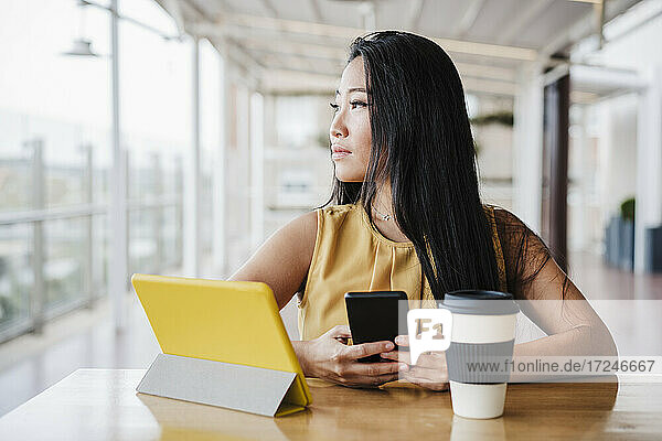 Unternehmerin schaut weg  während sie ihr Smartphone in einer modernen Büro-Cafeteria hält