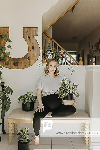 Schwangere Frau hält Topfpflanze  während sie auf einer Holzbank zu Hause sitzt