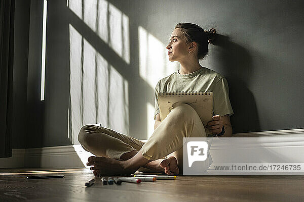 Young woman with note pad day dreaming while sitting on floor at home