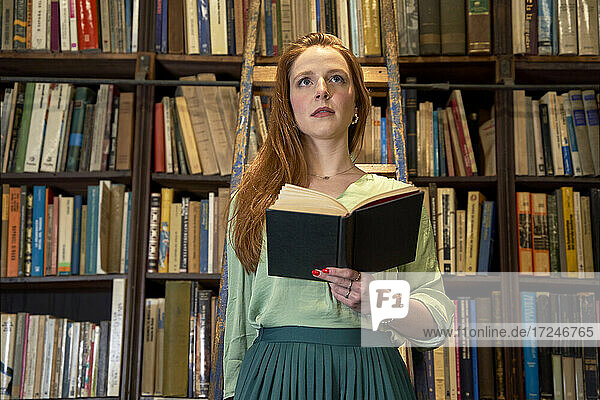 Junge Frau schaut weg  während sie auf einer Leiter in einer Bibliothek steht