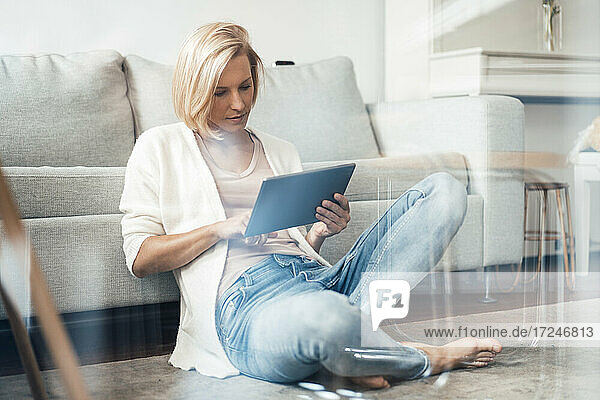 Blondhaarige Frau mit digitalem Tablet  die zu Hause auf dem Boden sitzt