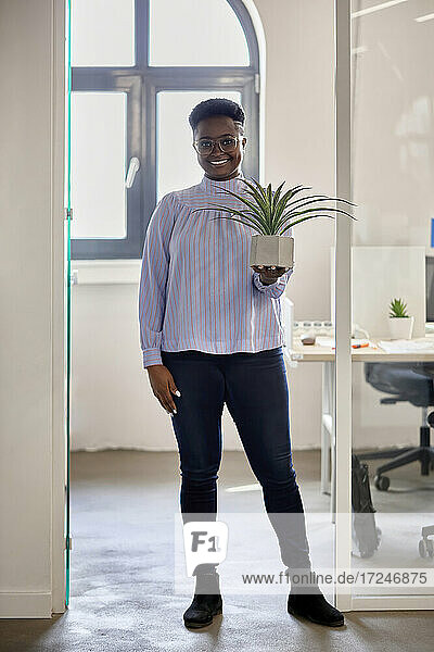 Lächelnde weibliche Fachkraft  die eine Pflanze hält  während sie in der Nähe der Eingangstür im Büro steht