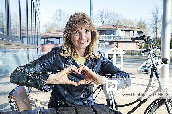 Mature woman showing heart shape sign while sitting by bicycle