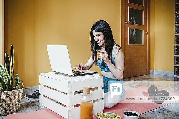 Weibliche Sportlerin  die einen Laptop benutzt  während sie zu Hause ein Trinkglas hält
