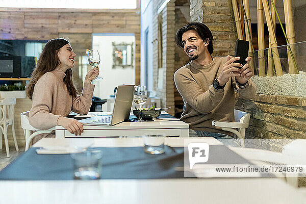 Lächelnder Mann  der ein Selfie mit seinem Smartphone macht  während er seine Freundin im Restaurant ansieht