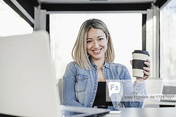 Glückliche Geschäftsfrau  die eine Einweg-Kaffeetasse hält  während sie mit einem Laptop in einem Café sitzt