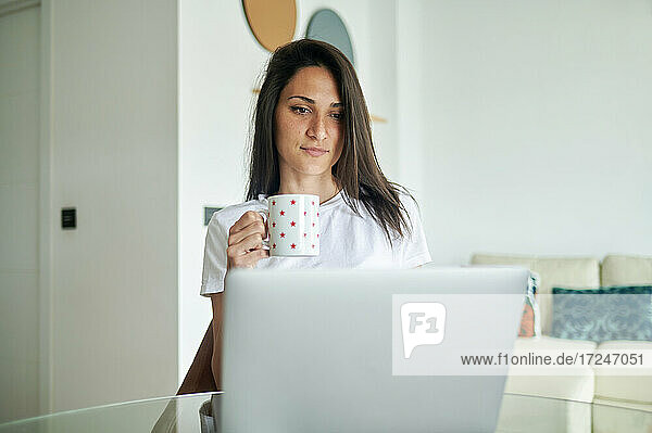 Mittlere erwachsene Frau hält eine Kaffeetasse  während sie zu Hause einen Laptop benutzt