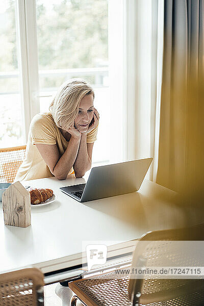 Berufstätige Frau mit Hand am Kinn  die zu Hause einen Laptop benutzt