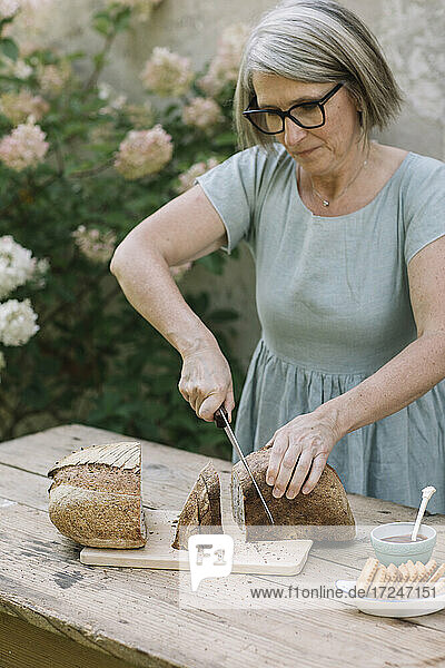 Reife Frau mit Brille schneidet Brot auf einem Tisch