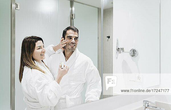 Lächelnde Frau schaut in den Spiegel  während sie Gesichtscreme auf das Gesicht eines Mannes im Hotelbad aufträgt