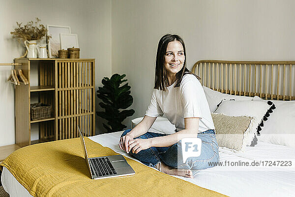 Lächelnde Frau sitzt mit Laptop auf dem Bett zu Hause