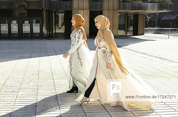 Modische Freundinnen mit Hijabs  die lächelnd auf dem Fußweg gehen