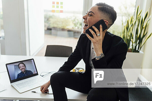 Glückliche Geschäftsfrau  die während eines Videogesprächs über einen Laptop im Büro ein Mobiltelefon benutzt