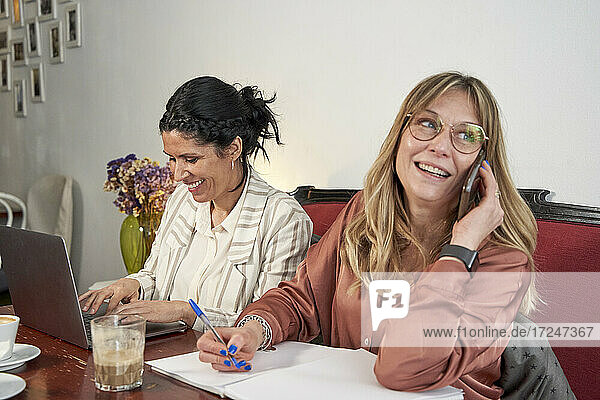 Lächelnde Geschäftsfrau  die wegschaut und mit einem Smartphone spricht  während sie mit einer Kollegin im Café sitzt