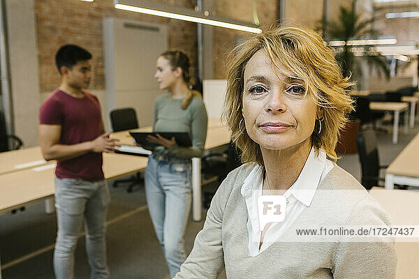 Reife Geschäftsfrau mit blondem Haar mit männlichen und weiblichen Mitarbeitern im Hintergrund im Büro