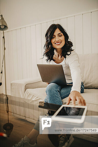 Freiberuflerin greift nach einem digitalen Tablet  während sie zu Hause am Laptop sitzt
