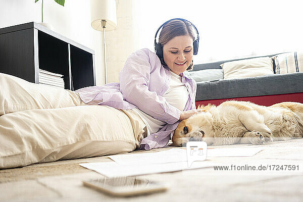 Lächelnde Frau mit Kopfhörern  die ihren Hund streichelt  während sie sich auf dem Teppich zu Hause entspannt