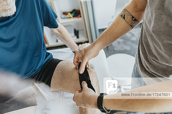 Male physiotherapist sticking elastic therapeutic tape on knee of patient