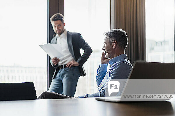 Lächelnder Geschäftsmann  der über sein Smartphone spricht  während sein Kollege im Büro an Dokumenten arbeitet