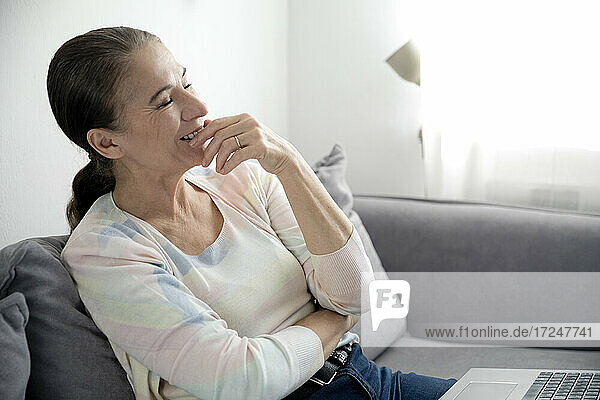Lächelnde Frau  die mit ihrem Laptop auf dem Sofa sitzt und träumt