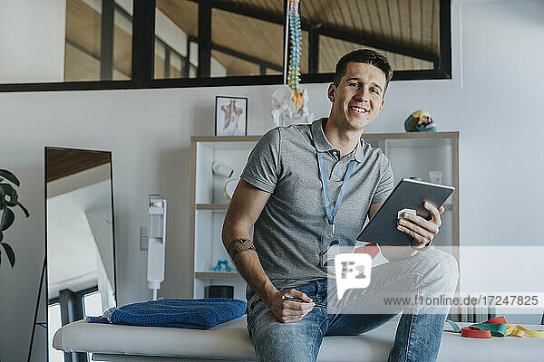 Lächelnder männlicher Physiotherapeut mit digitalem Tablet auf einem Bett in einer Arztpraxis sitzend
