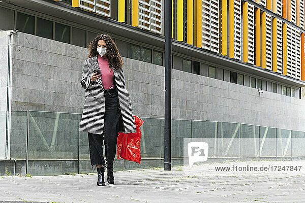 Junge Frau mit Jacke und Gesichtsmaske  die ein Mobiltelefon benutzt  während sie auf einem Fußweg geht