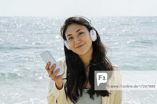 Lächelnde Frau mit Handy in der Hand  die an einem sonnigen Tag über Kopfhörer Musik hört
