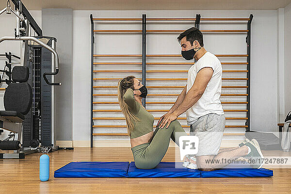 Male athlete with face mask helping female friend while exercising in health club