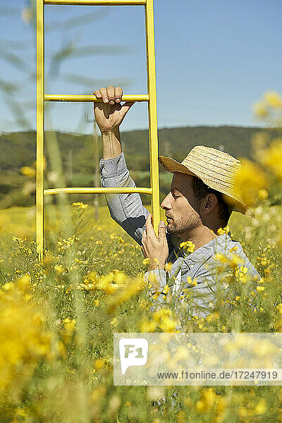 Ernster Mann mit Strohhut  der eine Leiter in einem Feld an einem sonnigen Tag hält