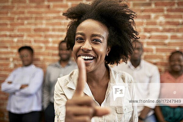 Fröhliche Geschäftsfrau zeigt Zeigefinger mit Kollegen im Hintergrund im Büro