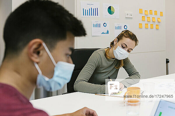 Männliche und weibliche Fachkraft mit Schutzmaske bei der Arbeit an einem Hot Desk in einem Coworking-Büro