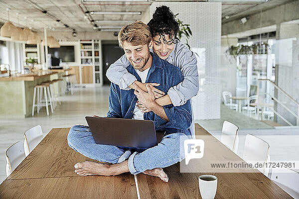 Lächelnde Frau umarmt Freund mit Laptop auf dem Tisch im Loft