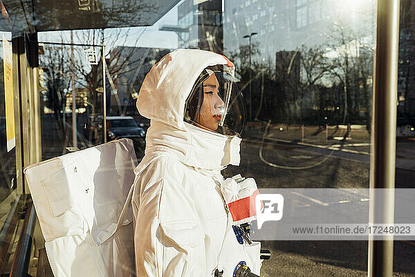 Weibliche Astronautin im Raumanzug an einer Bushaltestelle an einem sonnigen Tag durch Glas gesehen