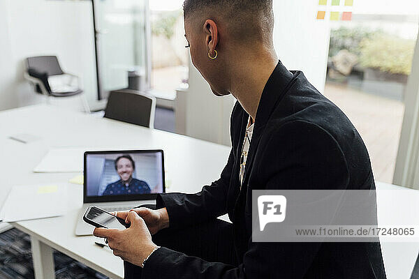Unternehmerin mit Mobiltelefon  die einen männlichen Kollegen bei einem Videogespräch über einen Laptop im Büro beobachtet