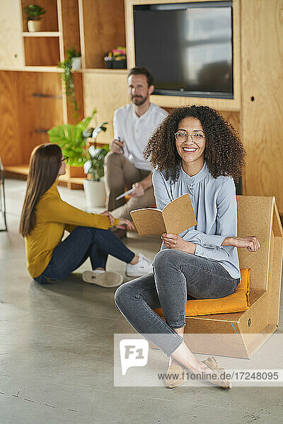 Smiling woman sitting on chair with colleagues in background at creative office