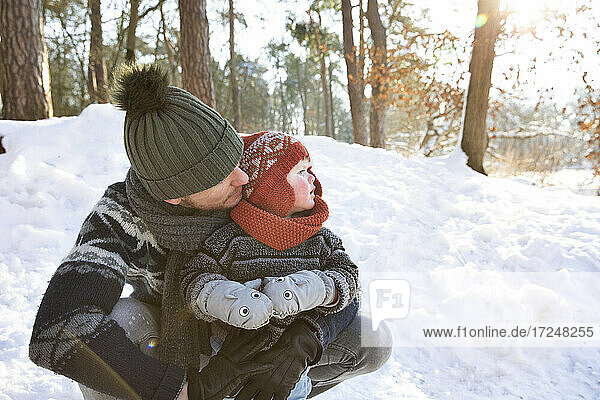 Father in knit hat embracing son while crouching on snow during winter