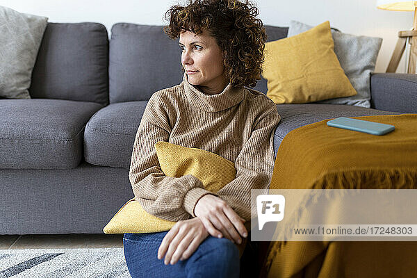 Mature woman looking away while sitting by sofa in living room at home