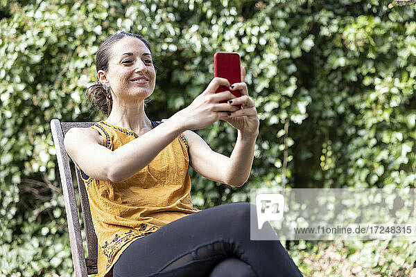 Lächelnde Frau  die ein Mobiltelefon benutzt  während sie auf einem Stuhl im Garten sitzt