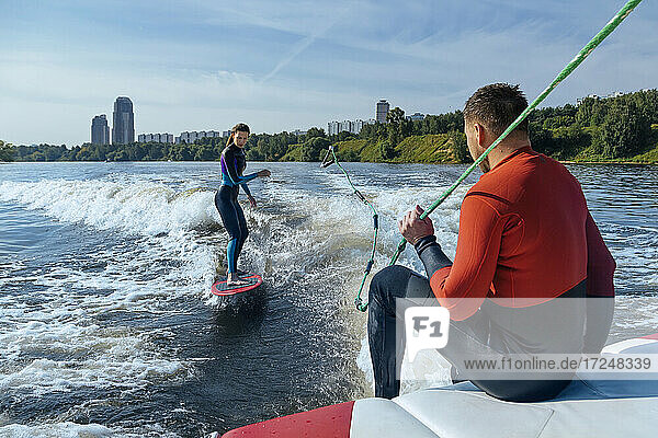 Mann sitzt am Rand eines fahrenden Bootes und schaut auf eine Wakeboarderin  die hinter ihm surft