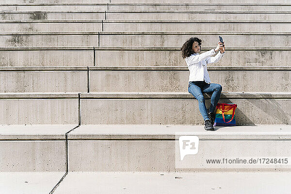 Hispanische Frau  die ein Selfie mit ihrem Smartphone macht  während sie an einem sonnigen Tag auf einer Treppe sitzt