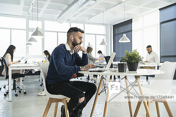 Male professional working on laptop with colleagues in background at coworking office