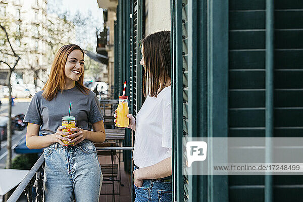 Smiling woman enjoying juice with girlfriend while standing at balcony