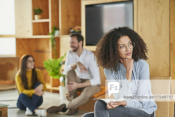 Nachdenkliche Geschäftsfrau im mittleren Erwachsenenalter  die wegschaut  während sie mit Kollegen im Hintergrund im Büro sitzt