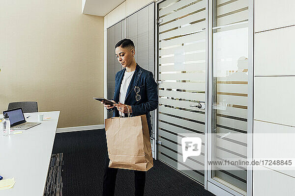 Geschäftsfrau mit digitalem Tablet im Stehen mit Papiertüte im Büro
