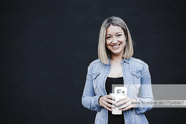 Geschäftsfrau lächelt  während sie eine Einweg-Kaffeetasse vor einer schwarzen Wand hält